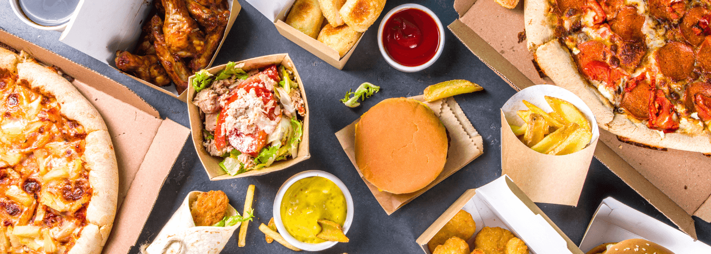 Overhead view of diverse takeout meal spread including pizza, burger, wings, fries, and salad in delivery containers