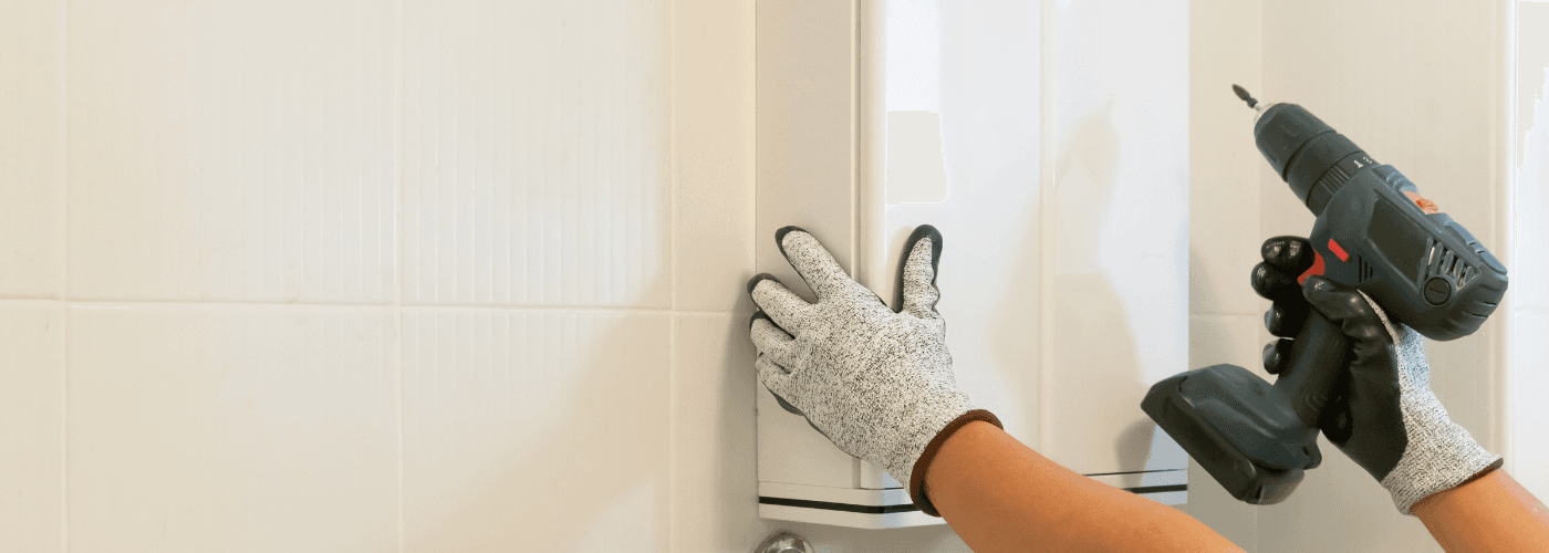 Hands wearing work gloves using cordless drill to install cabinet hardware on white kitchen cabinets