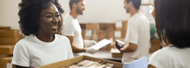 Smiling volunteer in white shirt holding donation box with team of volunteers sorting items in background