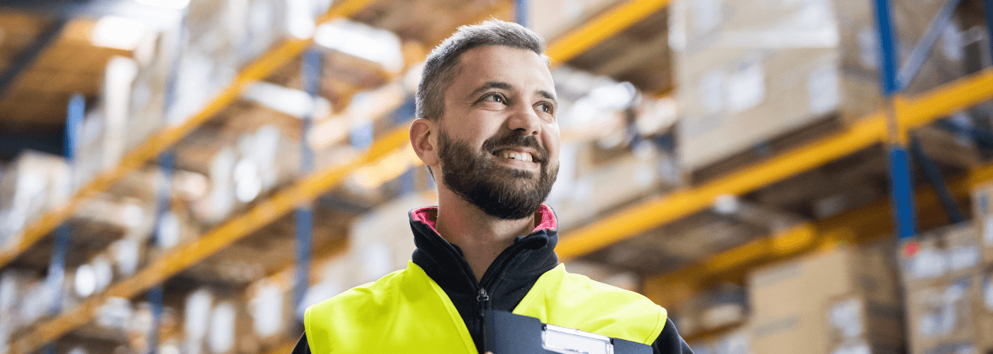 Smiling warehouse worker in safety vest standing in logistics facility with shelving racks of packages